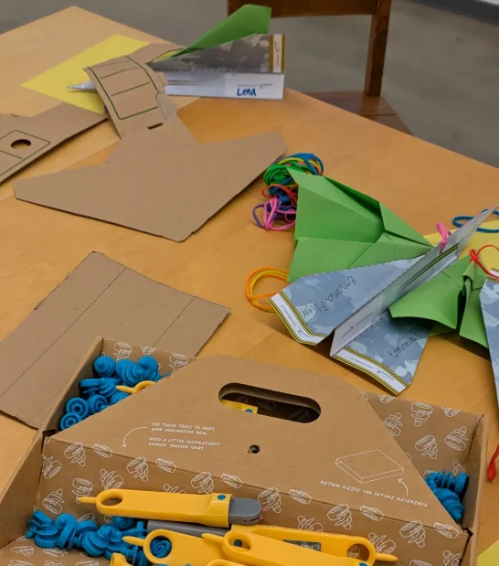 Was ist Auftrieb? Bunte Bastelmaterialien wie Papier, Scheren und Gummibänder auf einem Tisch.