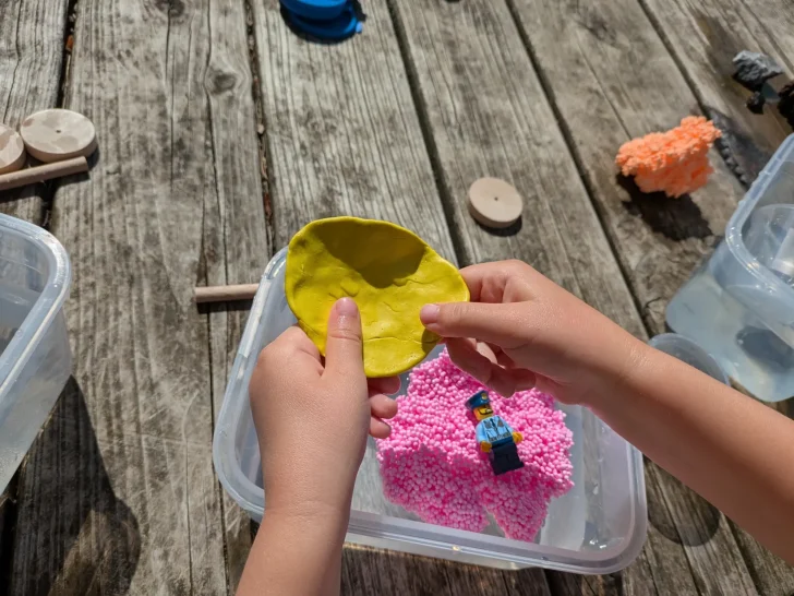 Schwimmt es? Zwei Hände formen gelbes Knetmaterial über pinkem Sand in einer Schüssel.