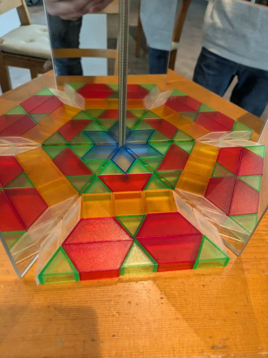 Spiegelexperiment Ein farbenfrohes geometrisches Muster aus roten, grünen und gelben Formen auf Holz.